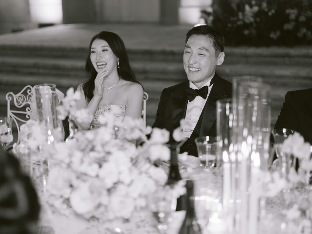 Casey and James laugh together behind soft flowers and glowing candles during their outdoor wedding dinner.