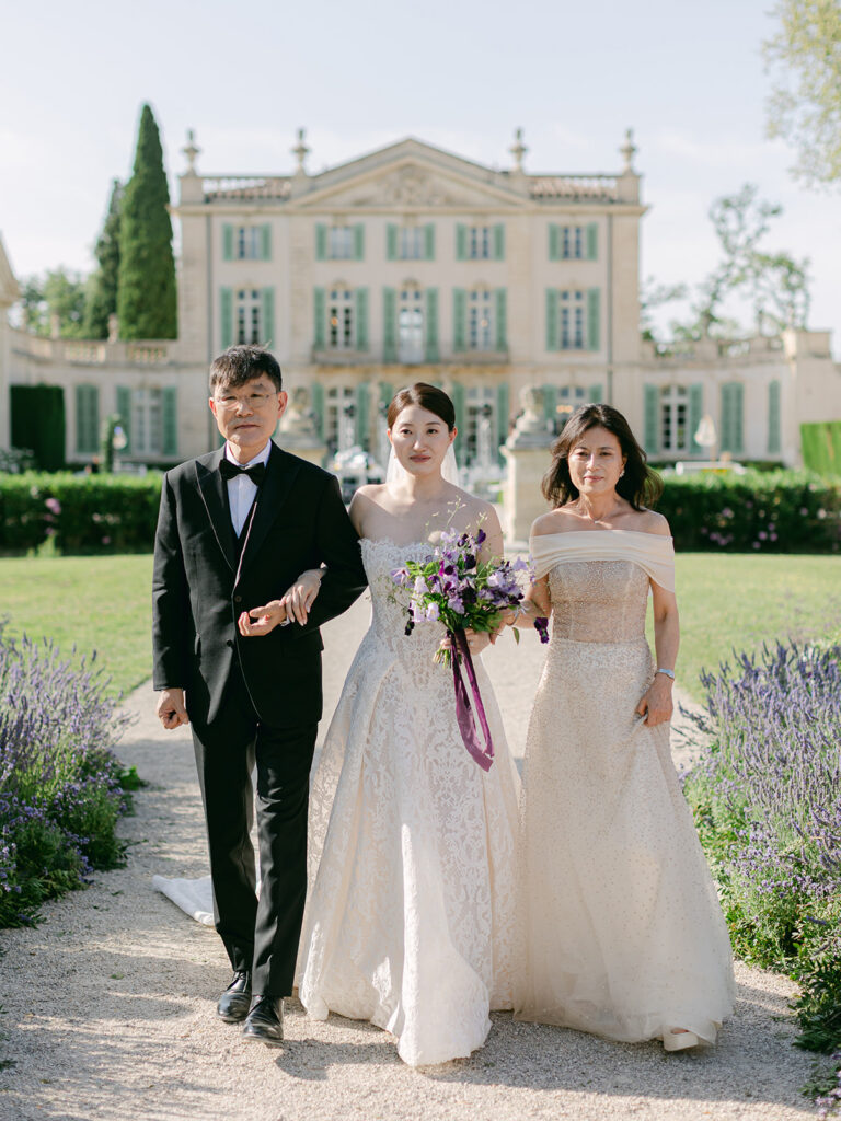 Casey walks with her parents holding a purple bridal bouquet before Château de Tourreau.