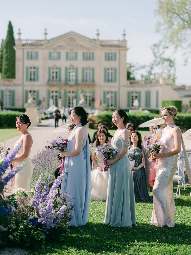 Bridesmaids with purple bouquets stand before Château de Tourreau during Casey and James's garden ceremony.