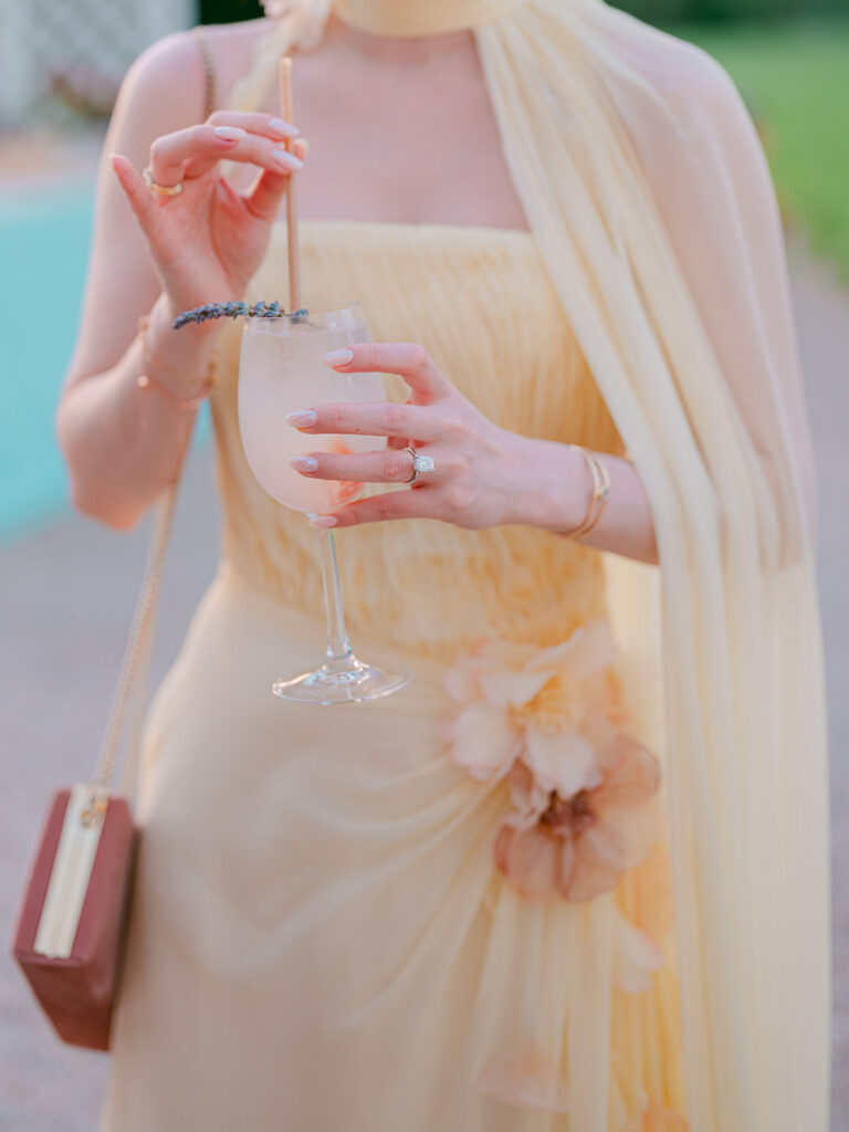 Close-up of Casey in a pale yellow dress holding a lavender-garnished cocktail beside the pool at Château de Tourreau.