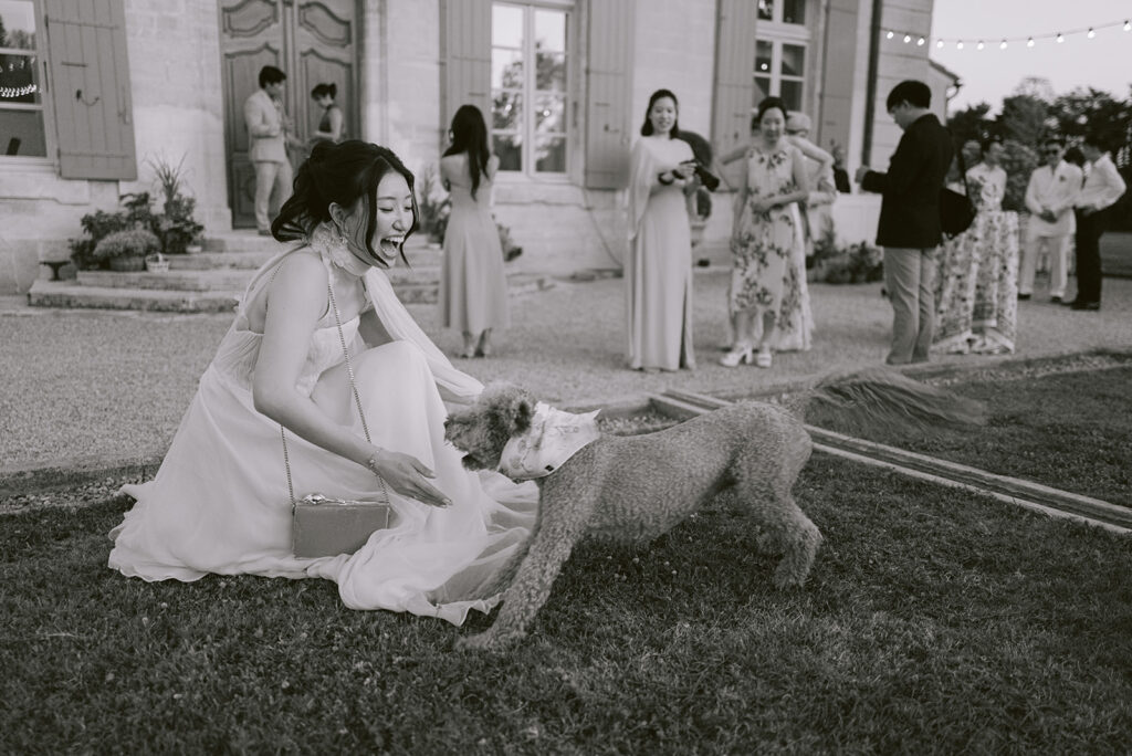 Black-and-white candid of Casey greeting the couple’s dog while guests mingle in the courtyard of Château de Tourreau.