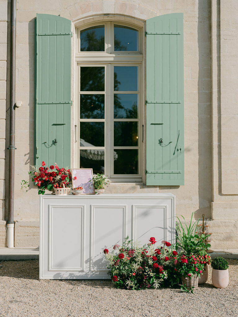 White cocktail bar decorated with red flowers, strawberry plants and a framed menu beneath the green shutters of Château de Tourreau.
