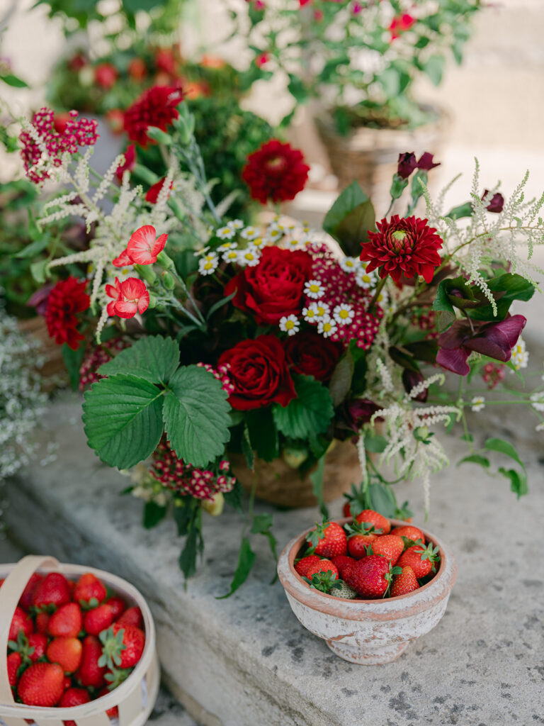 Red garden-style floral arrangement with roses, dahlias, astilbe and strawberry foliage beside bowls of fresh fruit.
