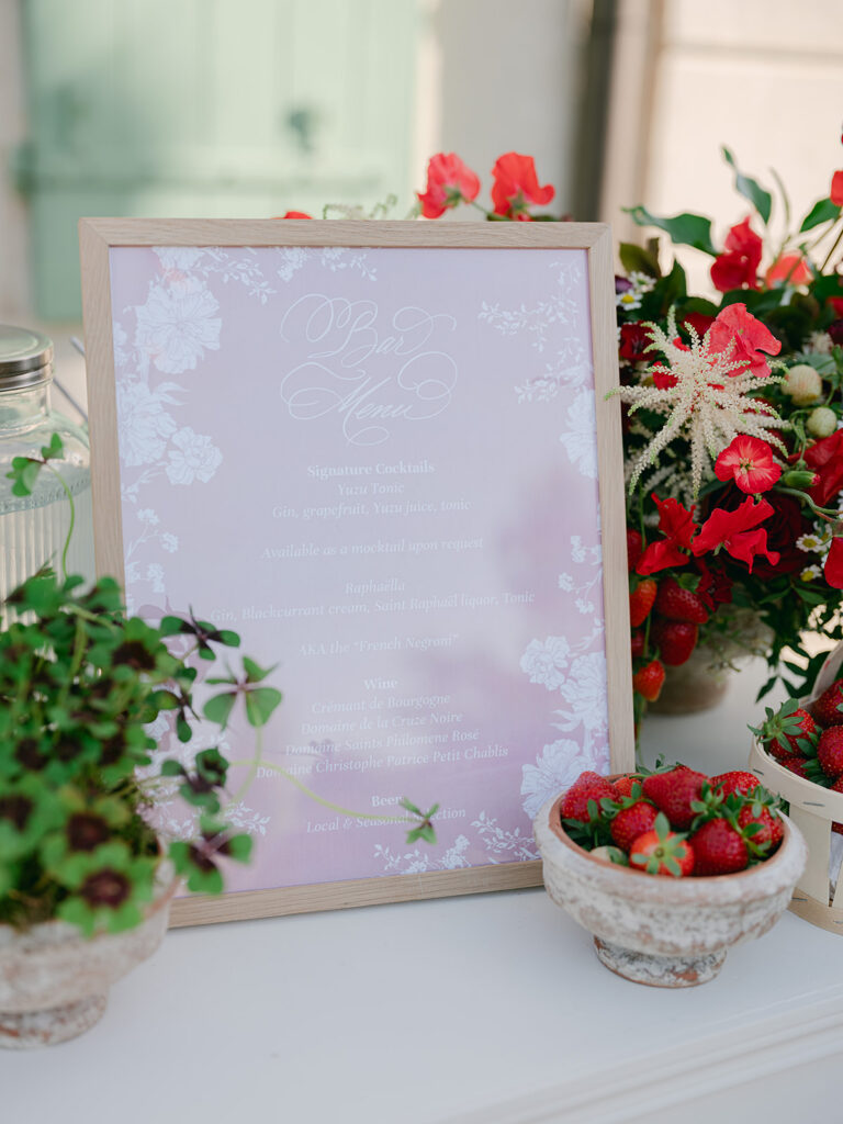 Framed signature cocktail menu surrounded by strawberries, oxalis and red garden flowers at Château de Tourreau.