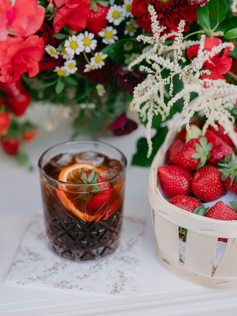 Dark cocktail garnished with citrus beside fresh strawberries and red summer flowers at the Château de Tourreau welcome event.