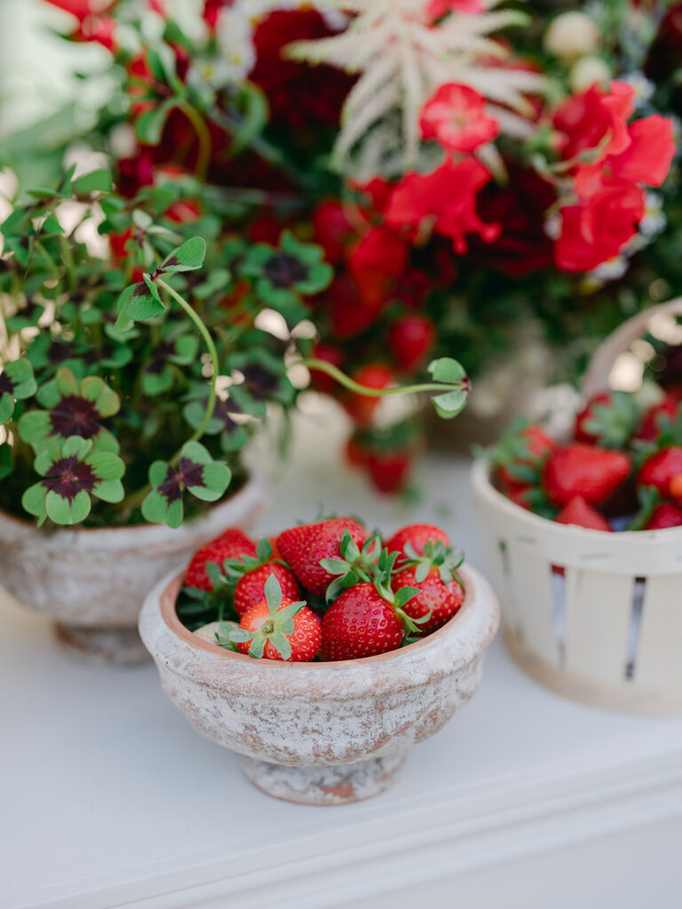 resh strawberries displayed in a terracotta bowl beside burgundy oxalis and a red garden-style floral arrangement.