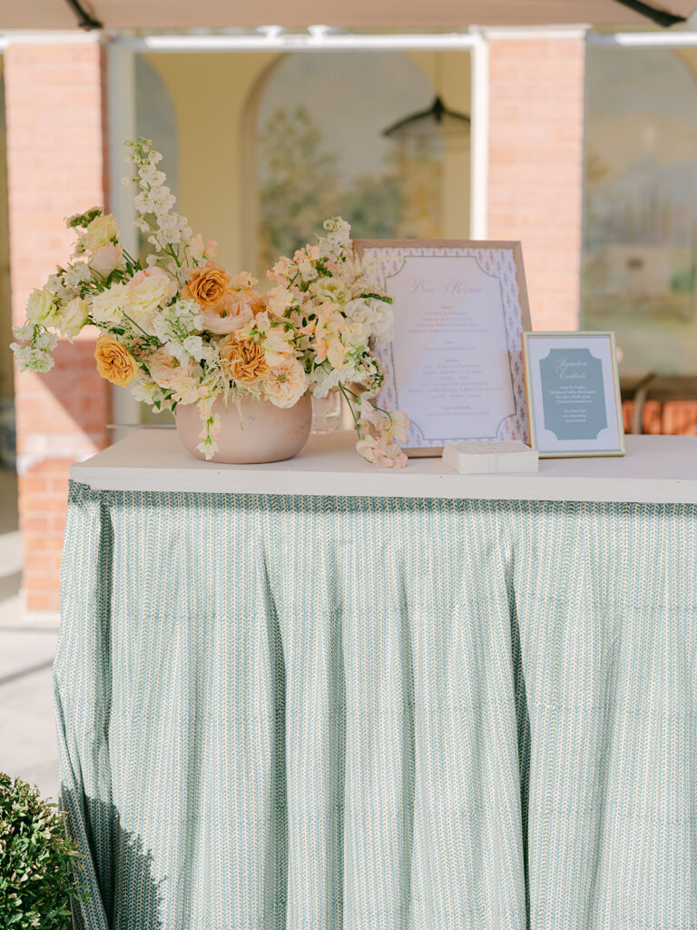 Sage-green wedding bar styled with peach floral arrangements and framed menus by Capucine Atelier Floral at Château de Tourreau.