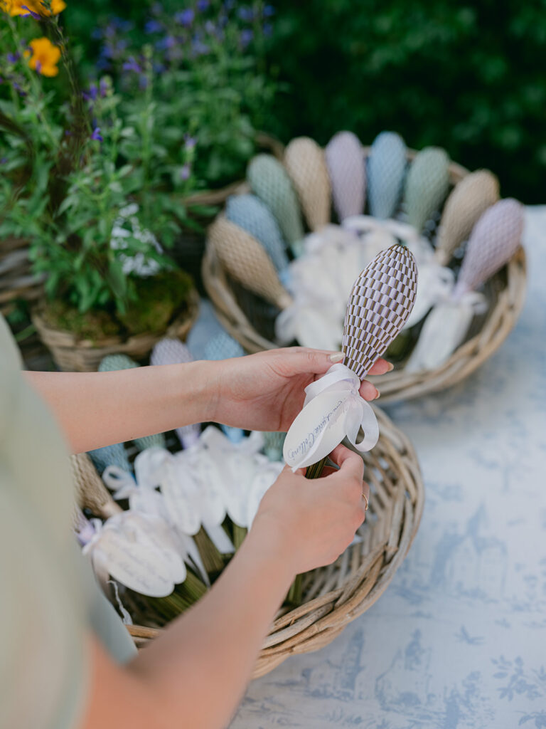 A guest selects a pastel woven fan with a white ribbon label from a wicker basket at the outdoor cocktail hour.