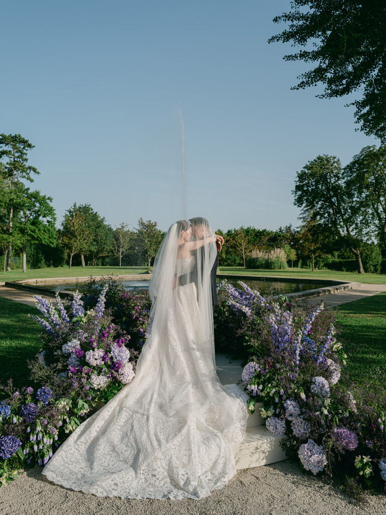 Newlyweds beneath the veil at the end of a flower-filled ceremony aisle overlooking the Château de Tourreau gardens.