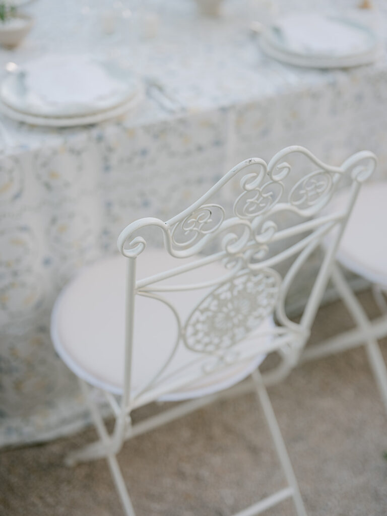 Ornate white wrought-iron dinner chair beside a softly patterned botanical tablecloth in the château courtyard.