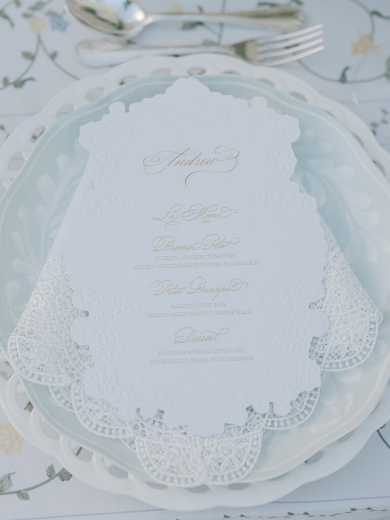 Personalized scalloped dinner menu with gold lettering displayed on elegant white wedding plates.