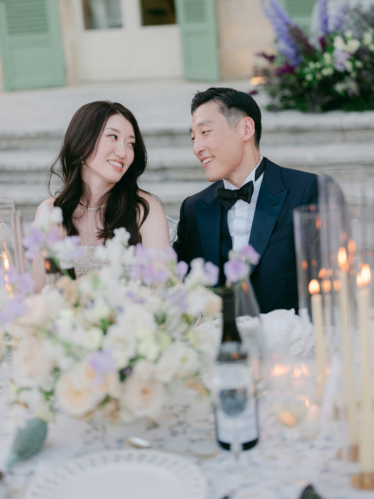 Wedding guests laugh together at a candlelit table framed by white, peach and lilac flowers.