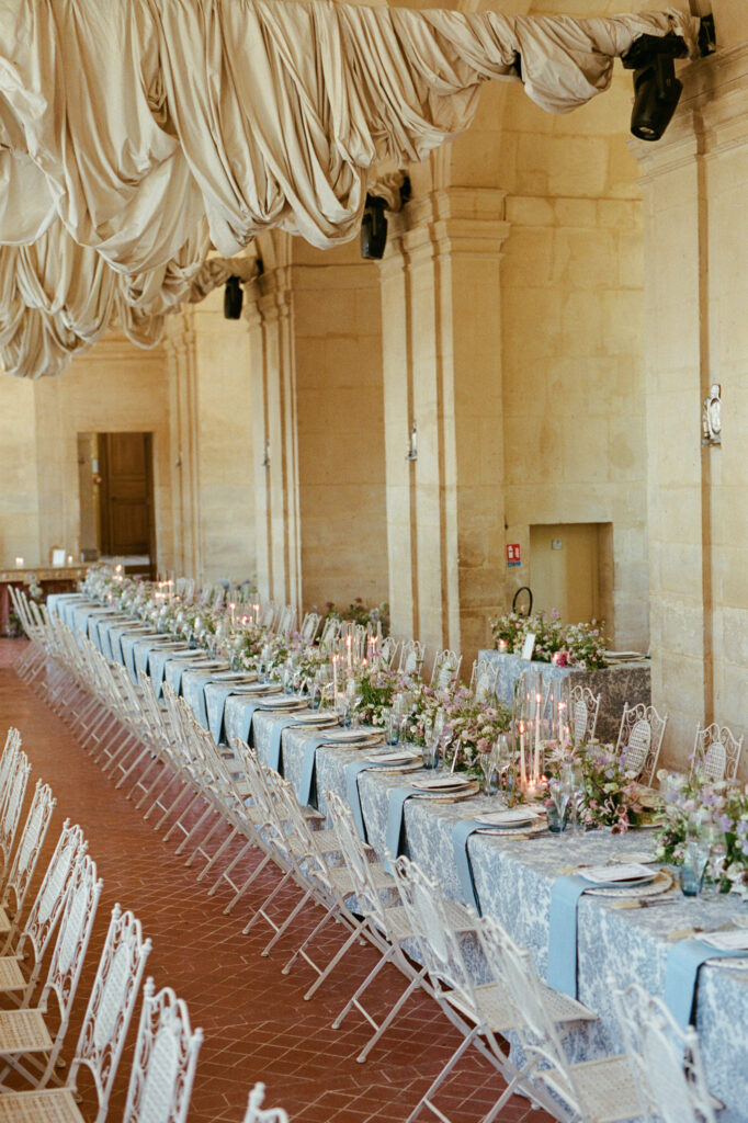 Rows of elegant banquet tables with blue linens and pastel flowers in the grand hall of Château de Vallery.