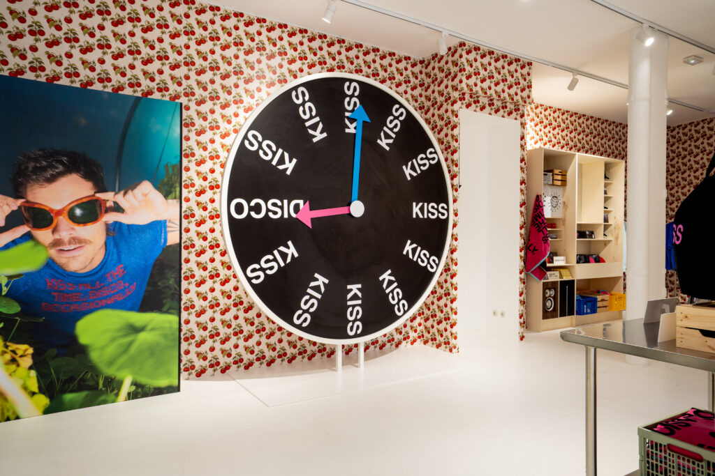 Oversized Kiss Disco clock beside a Harry Styles portrait and tomato-print walls at the Paris event.
