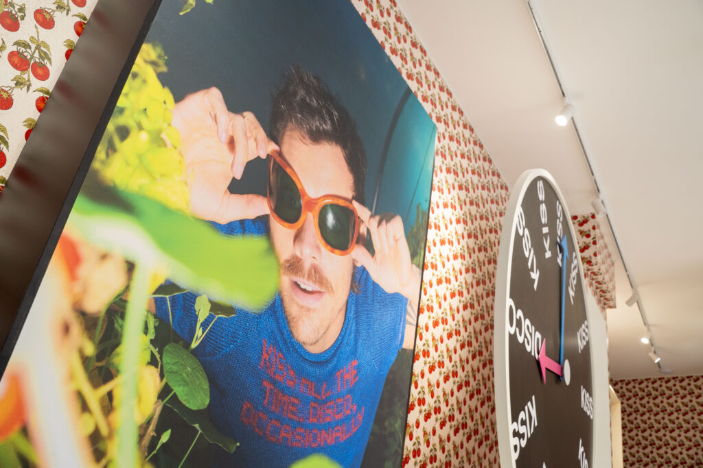 Oversized portrait of Harry Styles wearing orange sunglasses beside the Kiss Disco clock installation in Paris.