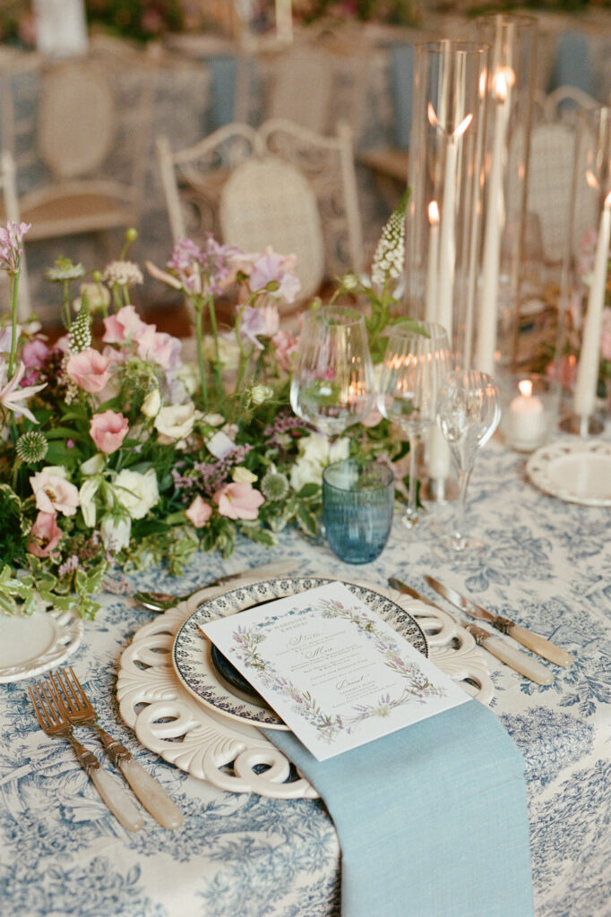 Layered place setting with pale blue napkin, illustrated menu, glass candlesticks and pastel garden flowers.
