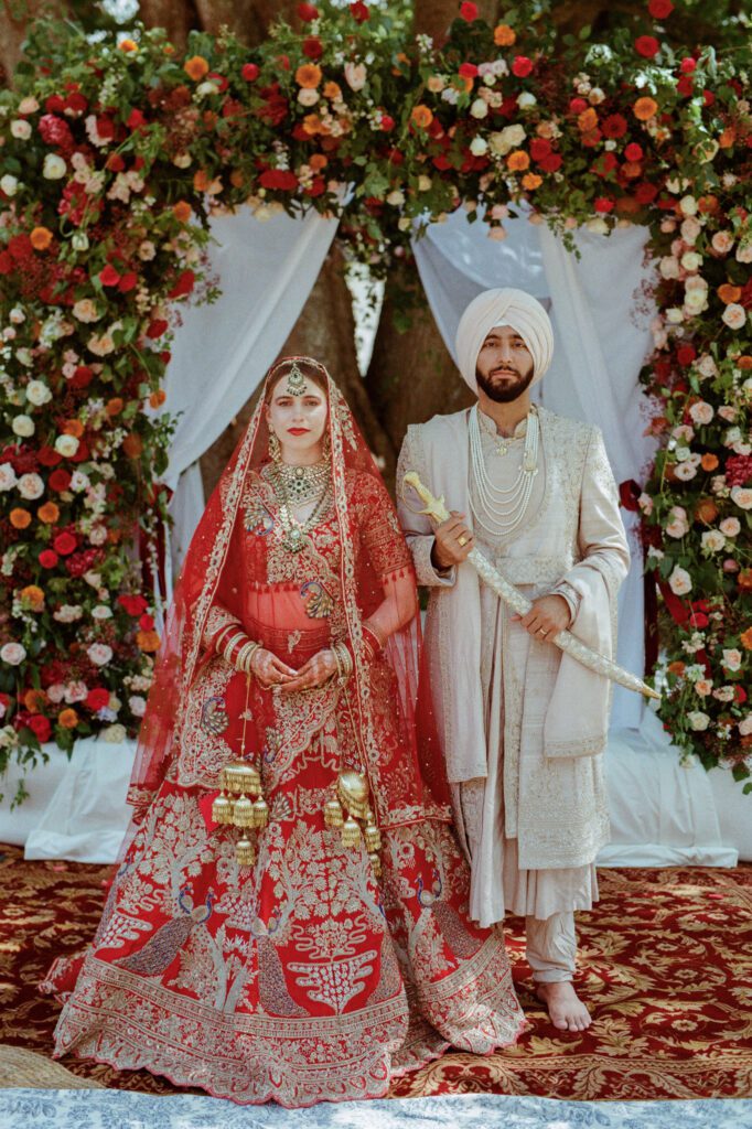 Kaylin in a red embroidered lehenga and Paul in an ivory sherwani pose before their abundant floral arch.