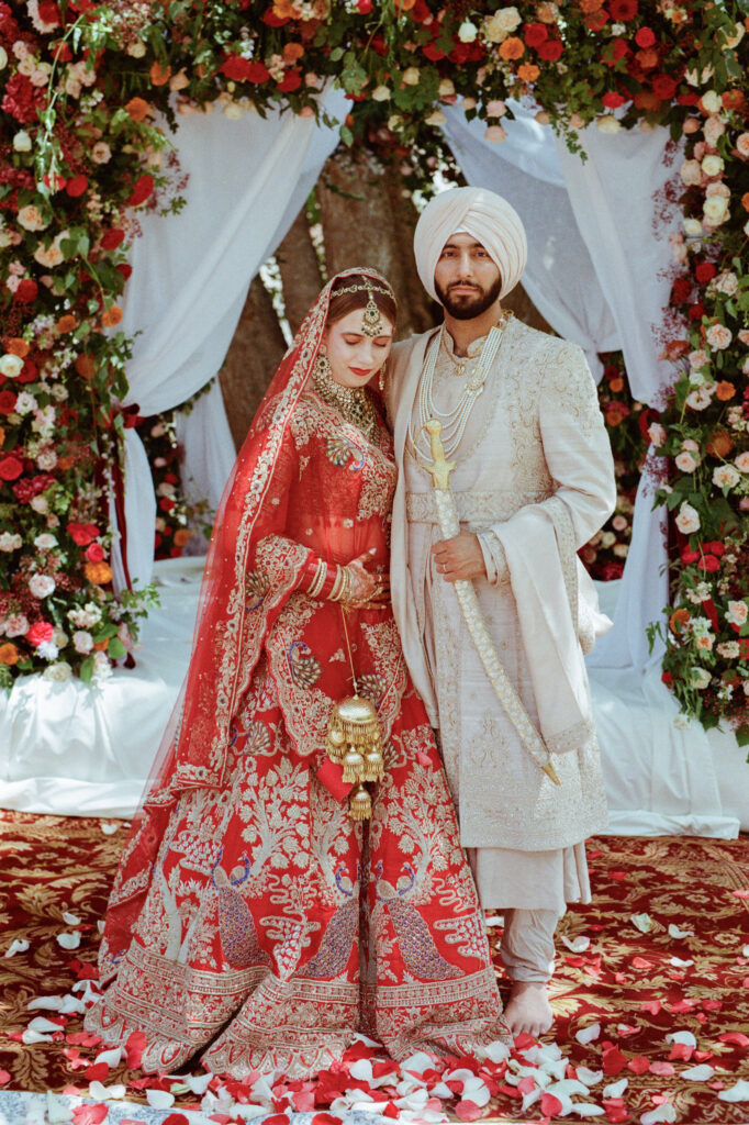 Kaylin in a red bridal lehenga sits beside Paul in an ivory sherwani during their Sikh wedding ceremony.