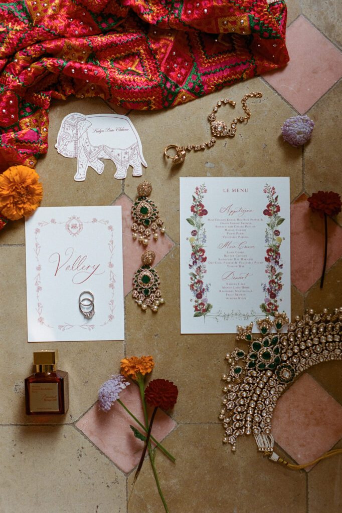 Indian wedding stationery, ornate jewelry, perfume and red embroidered fabric arranged in a fine-art flat lay.
