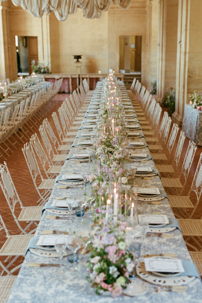 Long reception table with blue toile linen, pastel garden flowers and candles inside Château de Vallery.