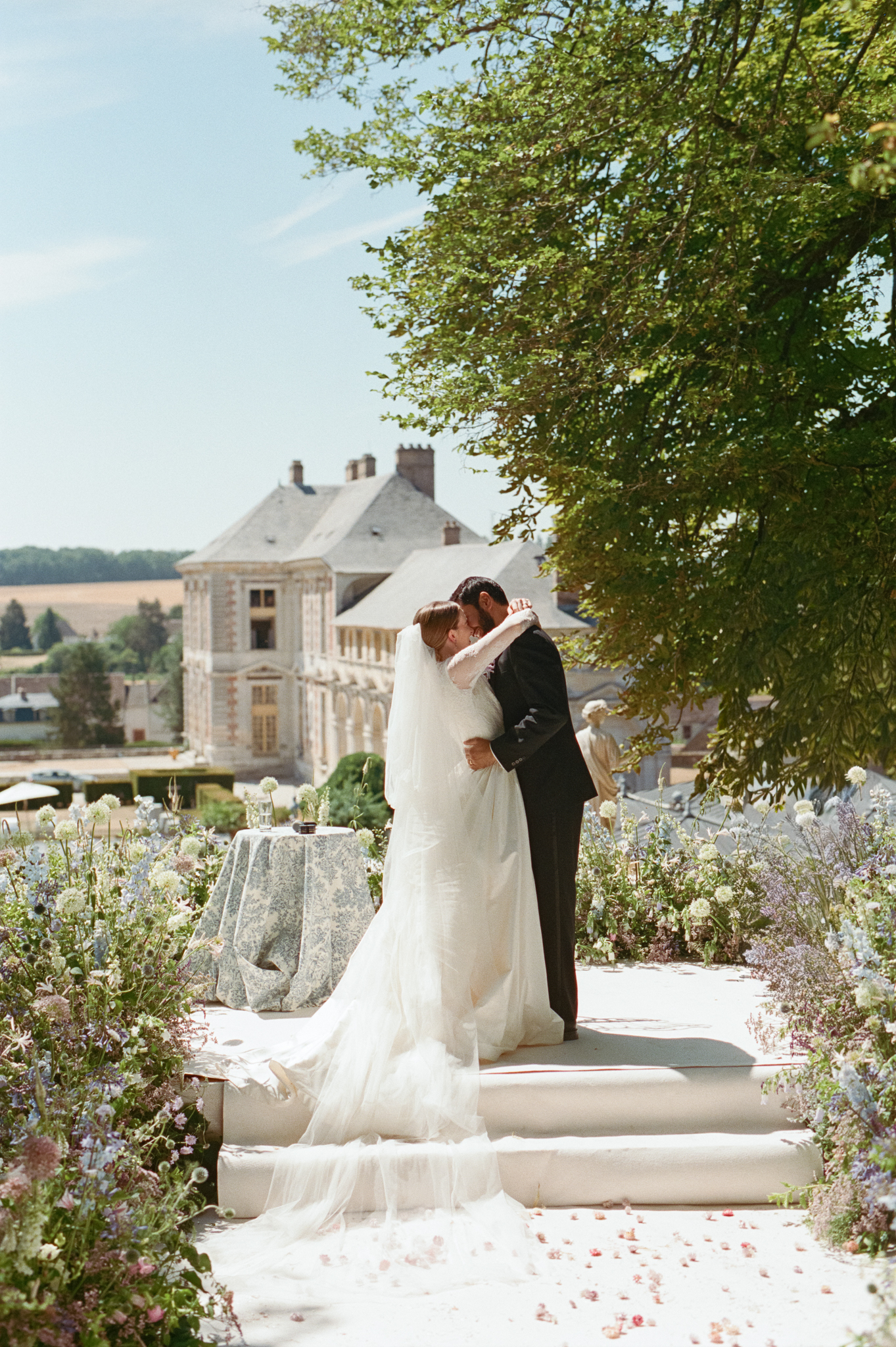 Kaylin and Paul kiss at the end of a flower-lined aisle with Château de Vallery in the background.