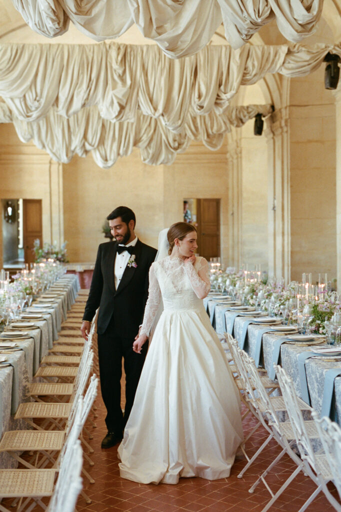 Kaylin and Paul walk between long flower-filled banquet tables inside Château de Vallery’s reception hall.