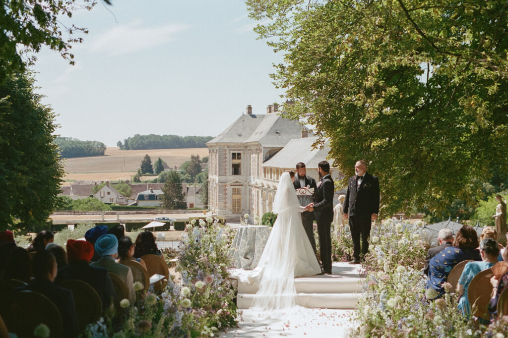 Kaylin and Paul exchange vows before guests along a pastel flower-lined aisle at Château de Vallery.