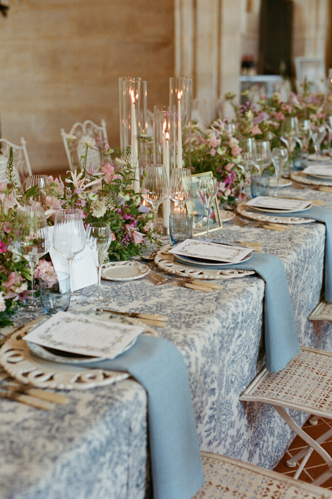 Long wedding table with blue toile linen, layered place settings, pastel flowers and slender glass candles.