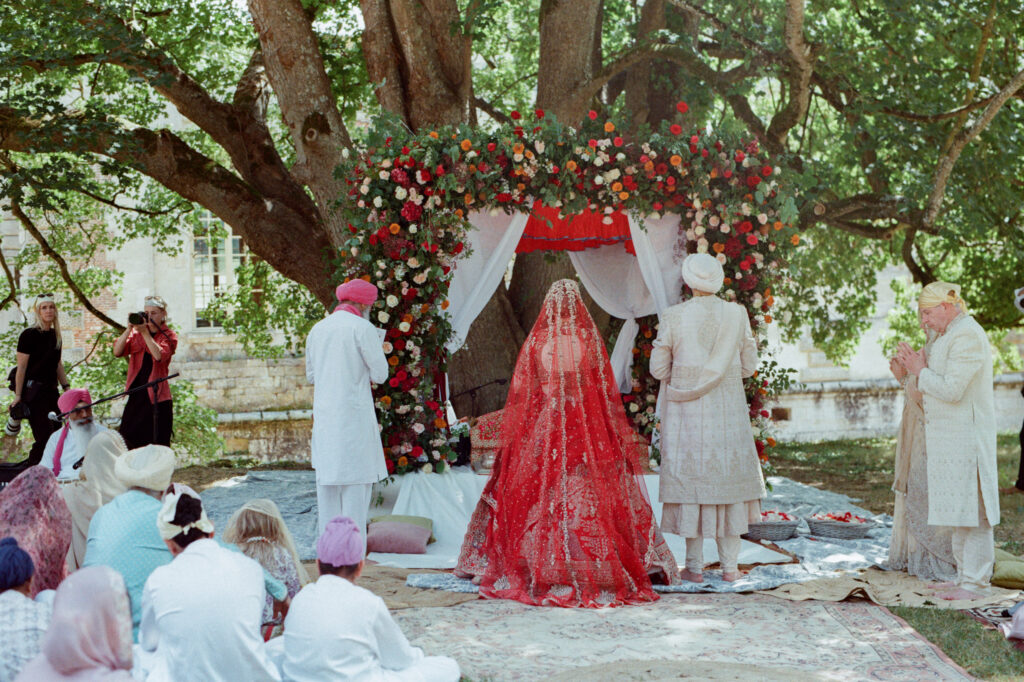 Kaylin and Paul beneath a red, orange and blush floral canopy by Capucine Atelier Floral during their Sikh ceremony.