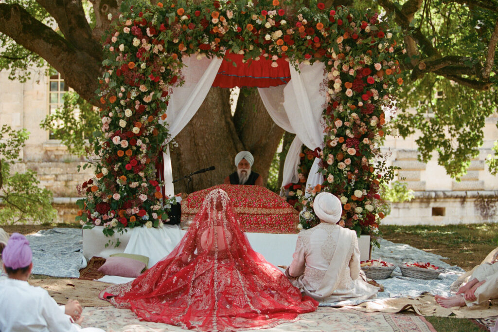 Kaylin and Paul sit before a richly flowered Sikh ceremony canopy beneath an old tree at Château de Vallery.